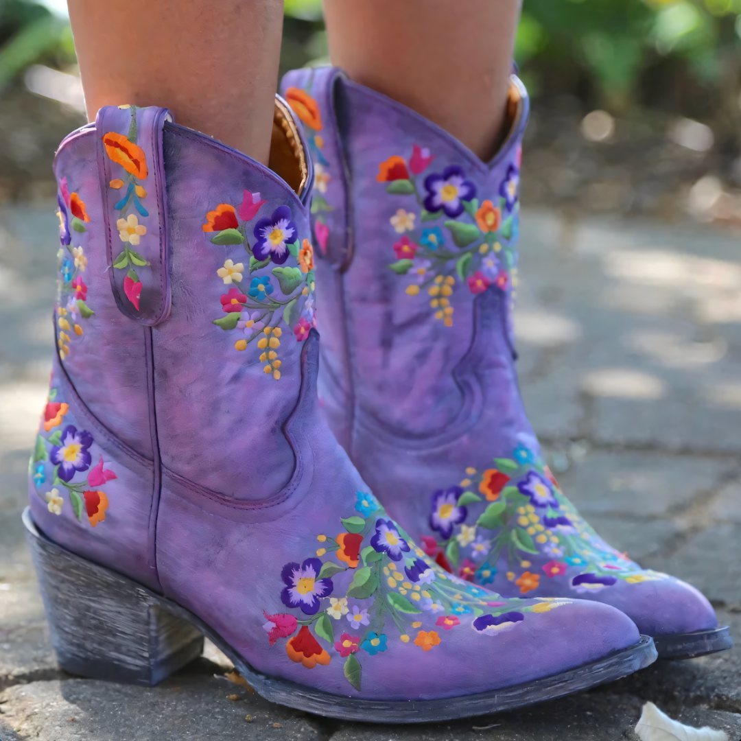 Paarse cowboylaarzen met kleurrijke bloemenborduursels, gedragen op een stenen pad. Trendy dameslaarzen, bohemian stijl, handgemaakt.