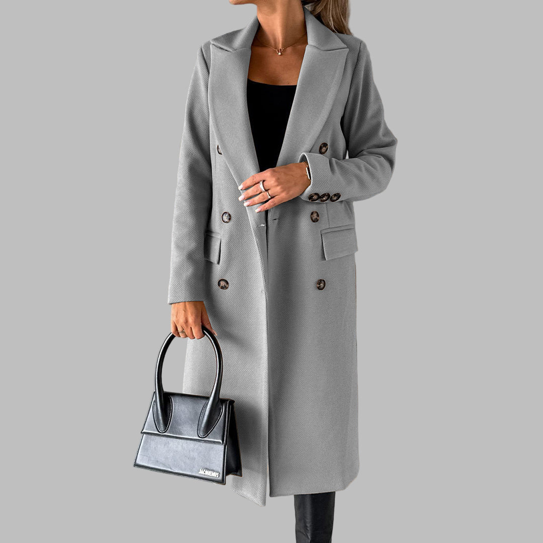 Grijze dames winterjas met dubbele knopenrij, gedragen met zwarte handtas en zwarte broek. Stijlvolle wintermode, elegante jas voor dames.