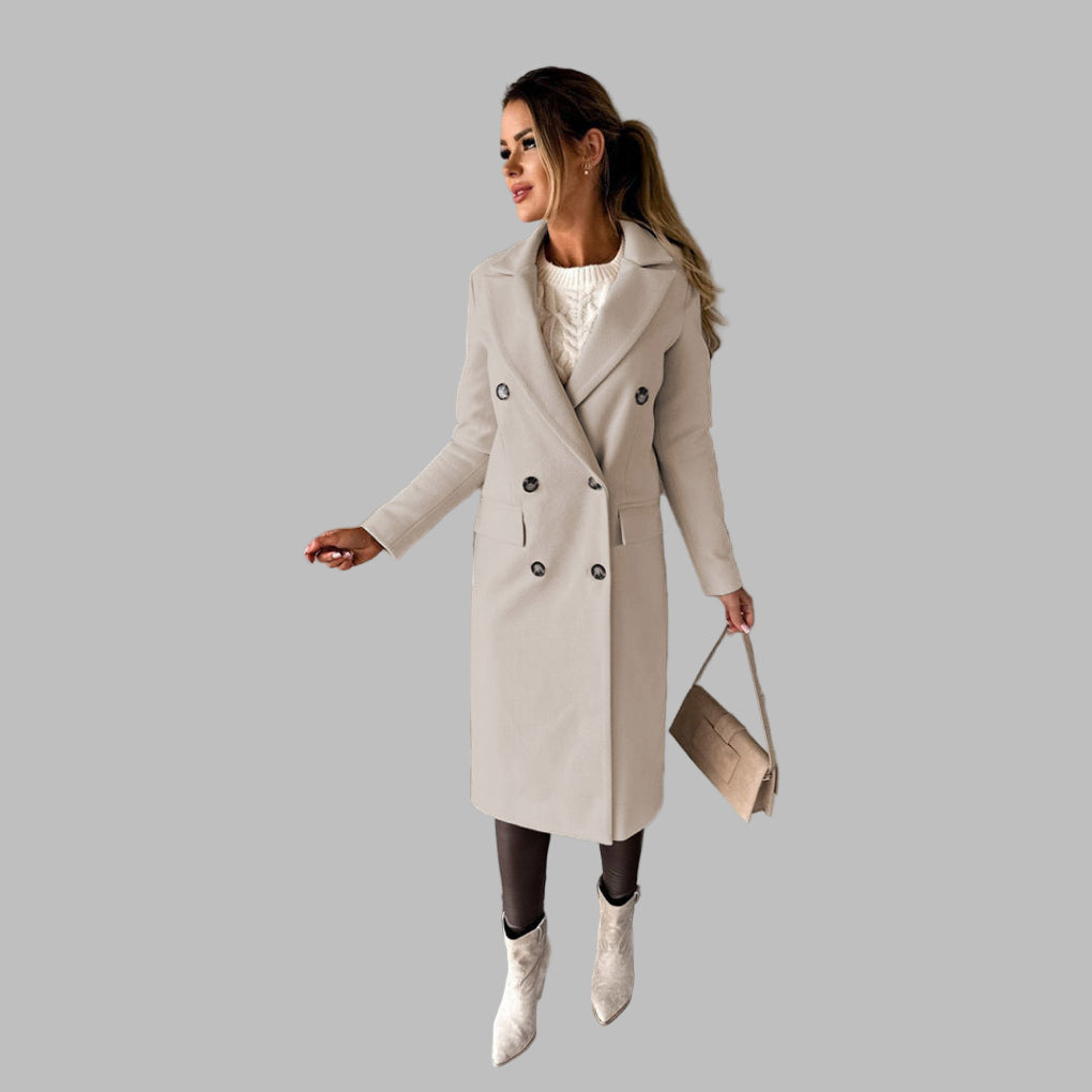 Vrouw in stijlvolle beige winterjas met dubbele rij knopen, witte trui en laarzen, poseert tegen een betonnen muur. Wintermode, dameskleding.